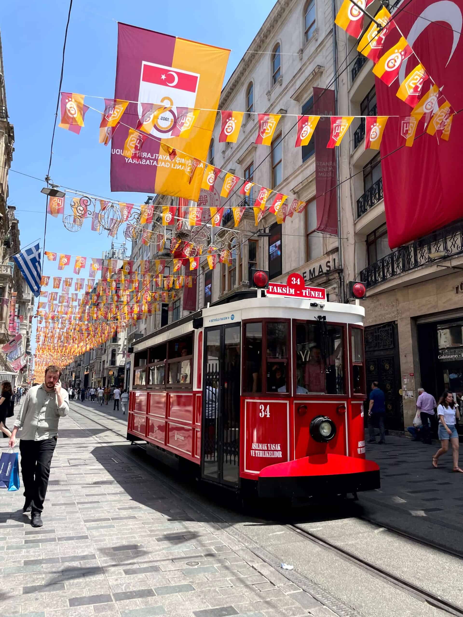 Historic red tram number 34 travels along İstiklal Street beneath red and yellow Galatasaray flags. A large Turkish flag hangs from a building beside the tracks.