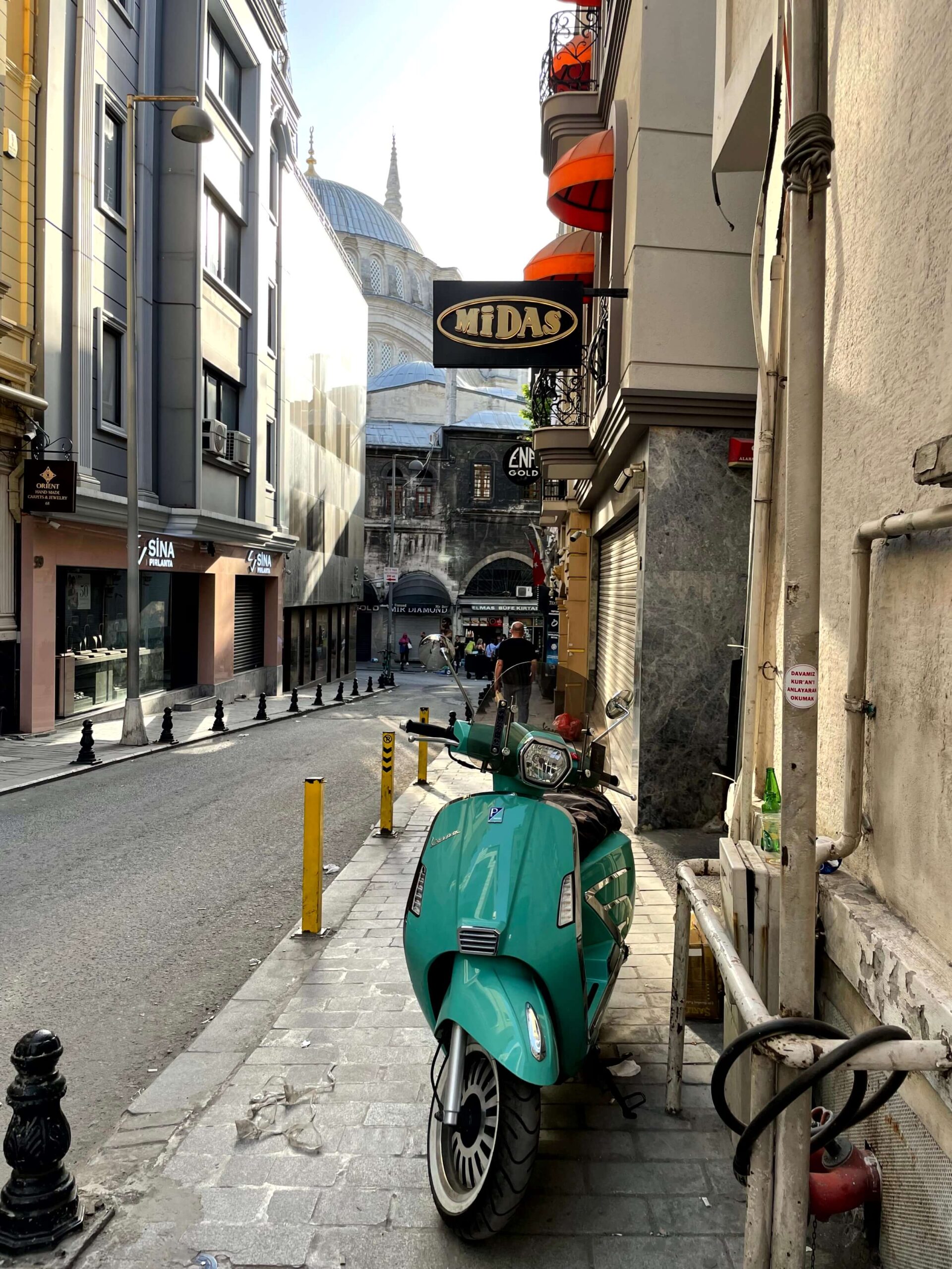 A turquoise vespa parked on a narrow Sultanahmet sidewalk with shop signs and a domed mosque in the background. The quiet street is lined with tall buildings and bollards.