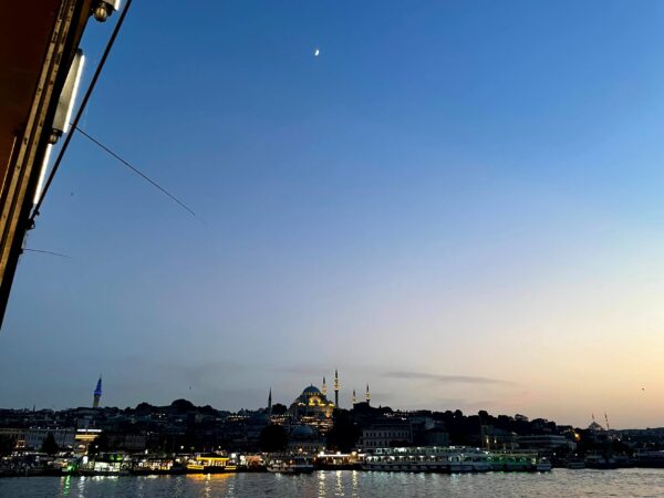 The view from Galata Bridge at dusk with Istanbul’s waterfront lit along the water. A mosque with glowing minarets rises on the skyline beneath a deep blue sky and small crescent moon.