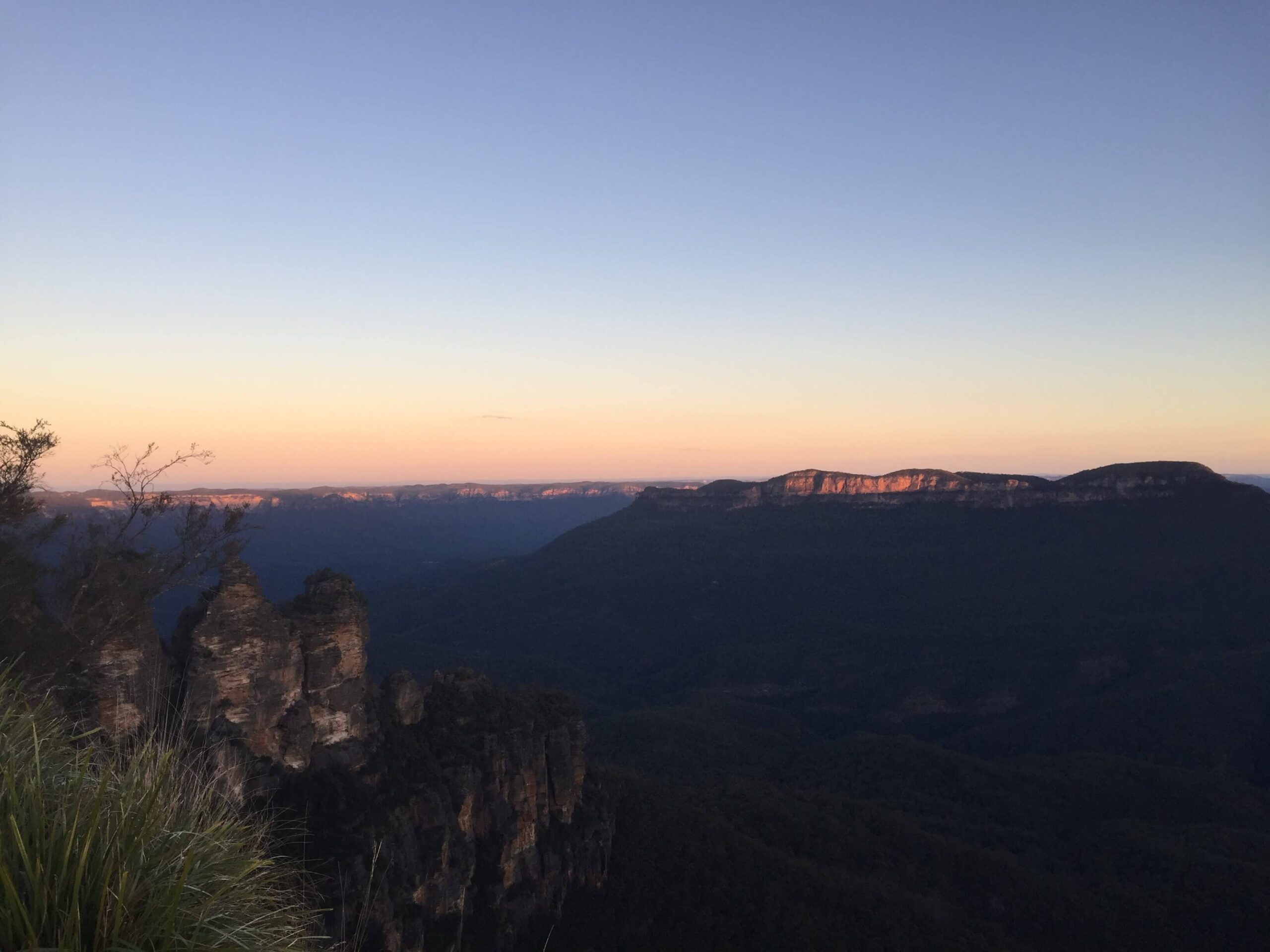 The Three Sisters rock formation viewed from Echo Point Lookout in the Blue Mountains at sunset with layered cliffs in the distance.