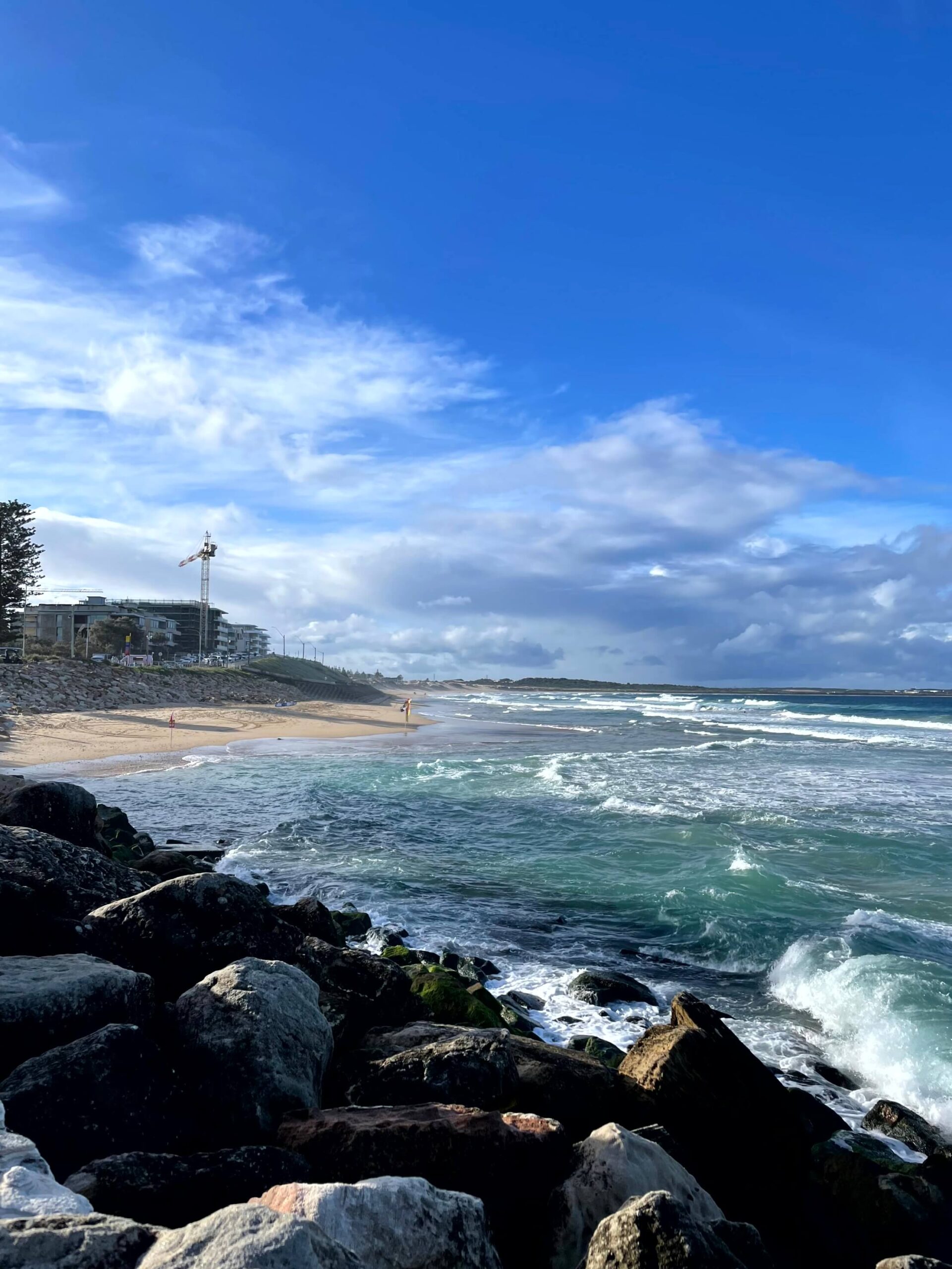 Waves crashing onto North Cronulla beach with rocks in the foreground under a bright blue sky with some clouds.