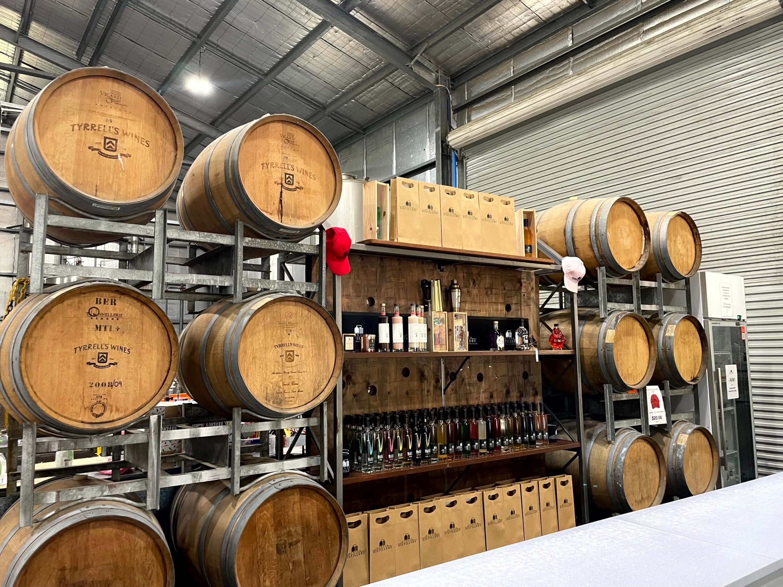 Rows of large wooden wine barrels stacked inside a winery in the Hunter Valley with bottles and boxes displayed on shelves.