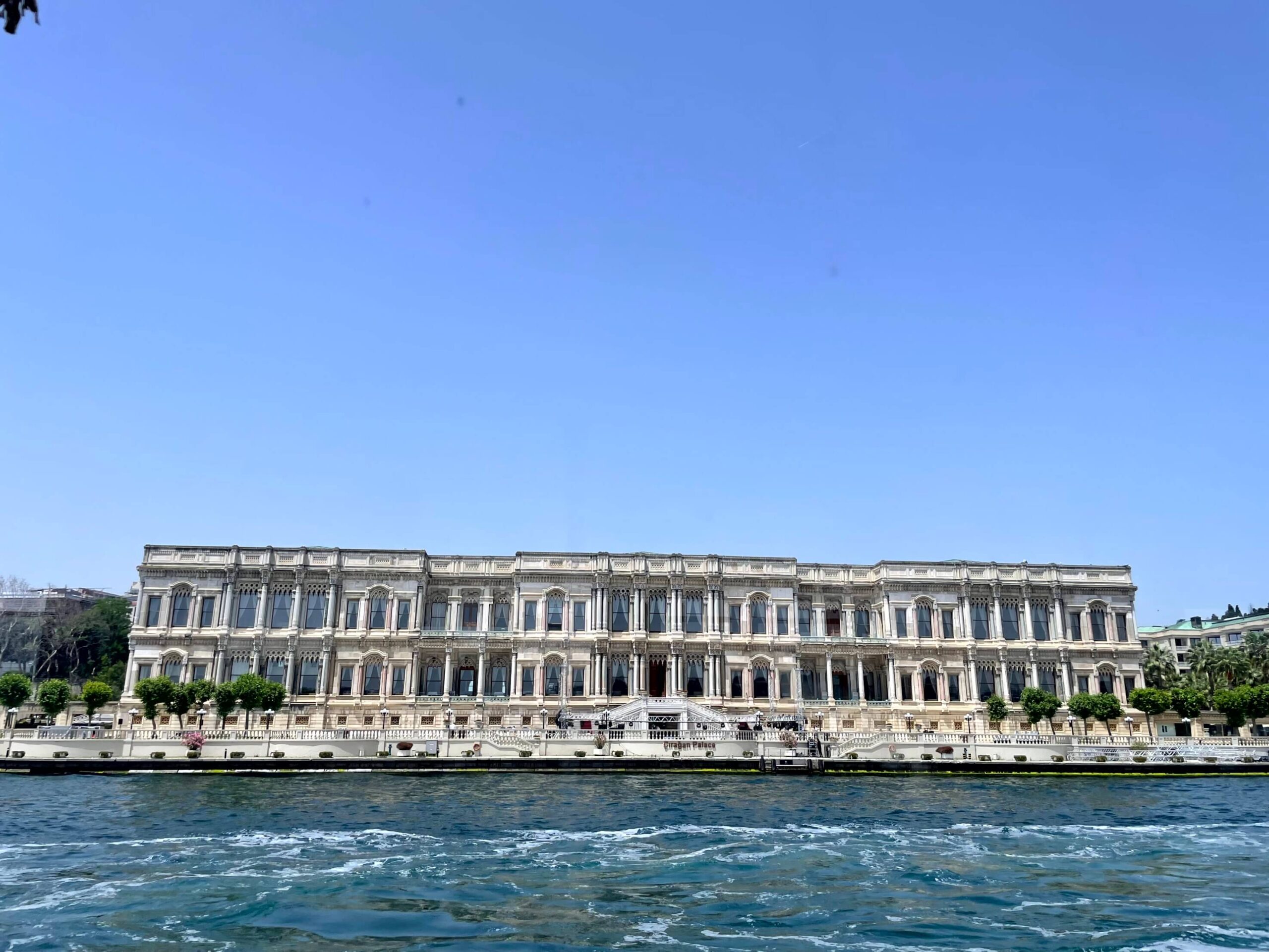 Ciragan Palace Kempinski stretches along the Bosphorus waterfront under a clear blue sky. Choppy water fills the foreground as the palace is seen from the water.