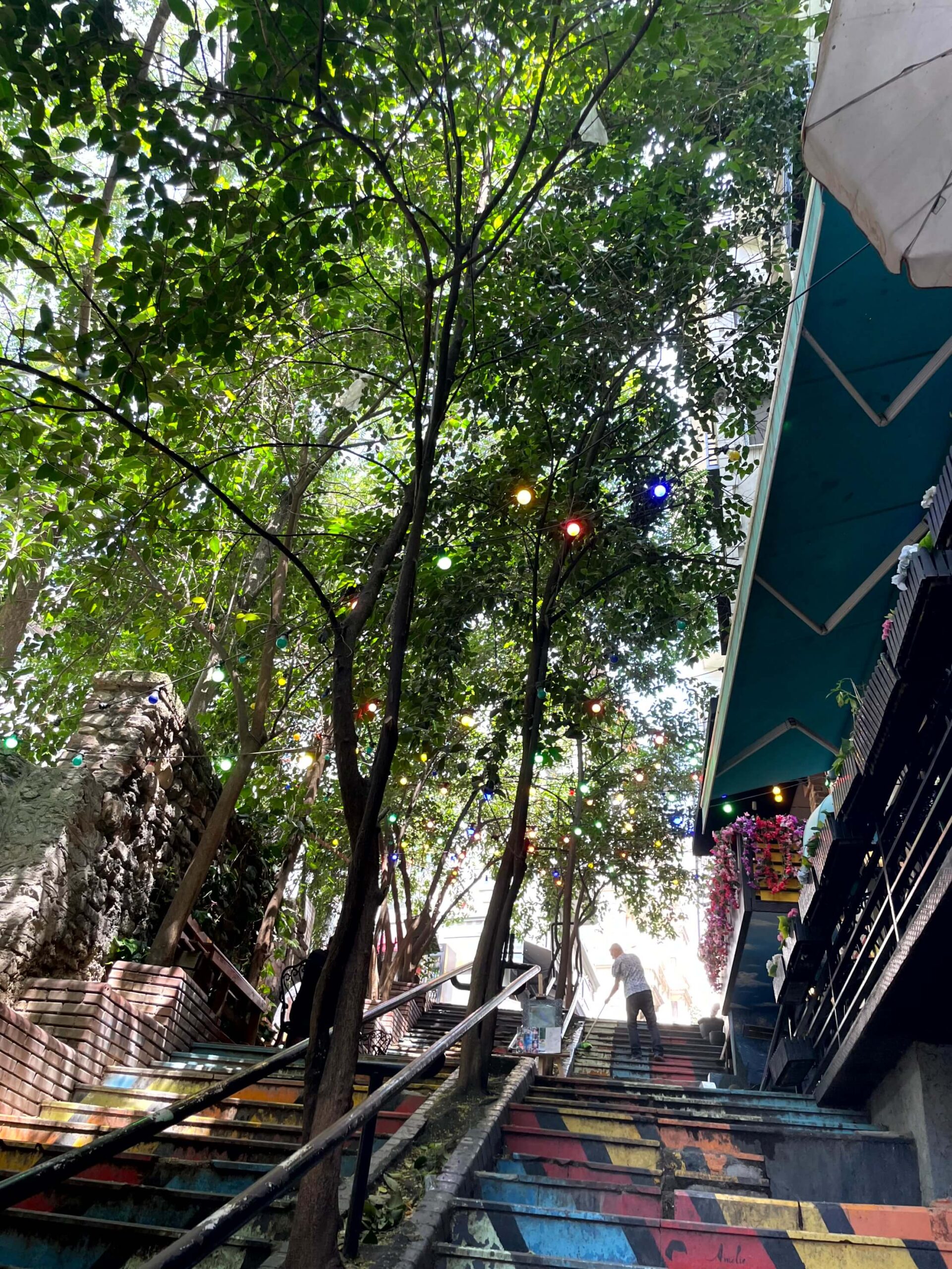 Colourful painted stairs in Beyoğlu shaded by green trees with string lights hanging overhead. A person stands near the top of the steps beside a building with bright flowers on the balcony.