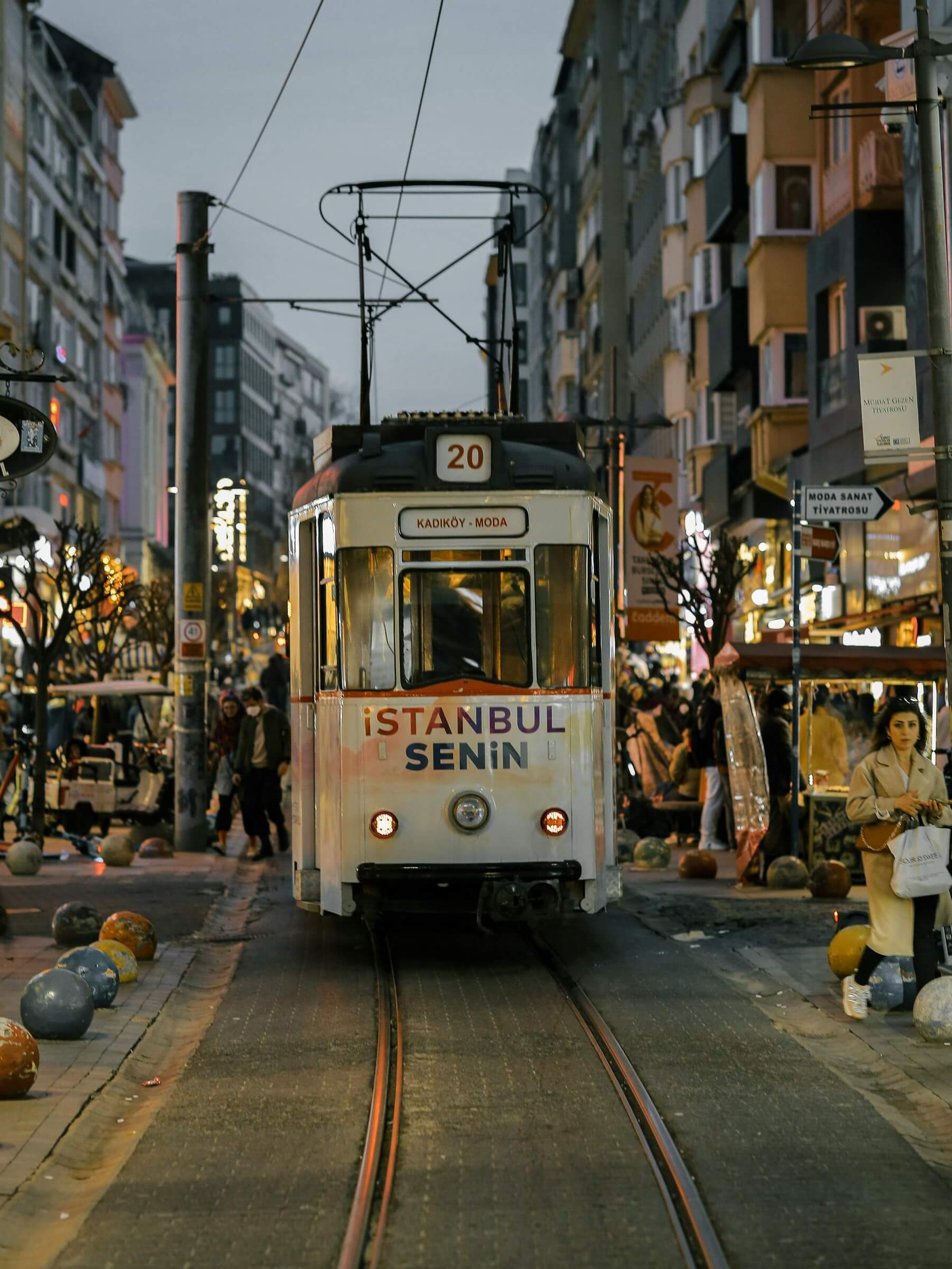 Vintage tram number 20 travels through Caferağa at dusk with headlights on. The tram reads "Kadıköy - Moda" and "İstanbul Senin" as people walk along the busy street.