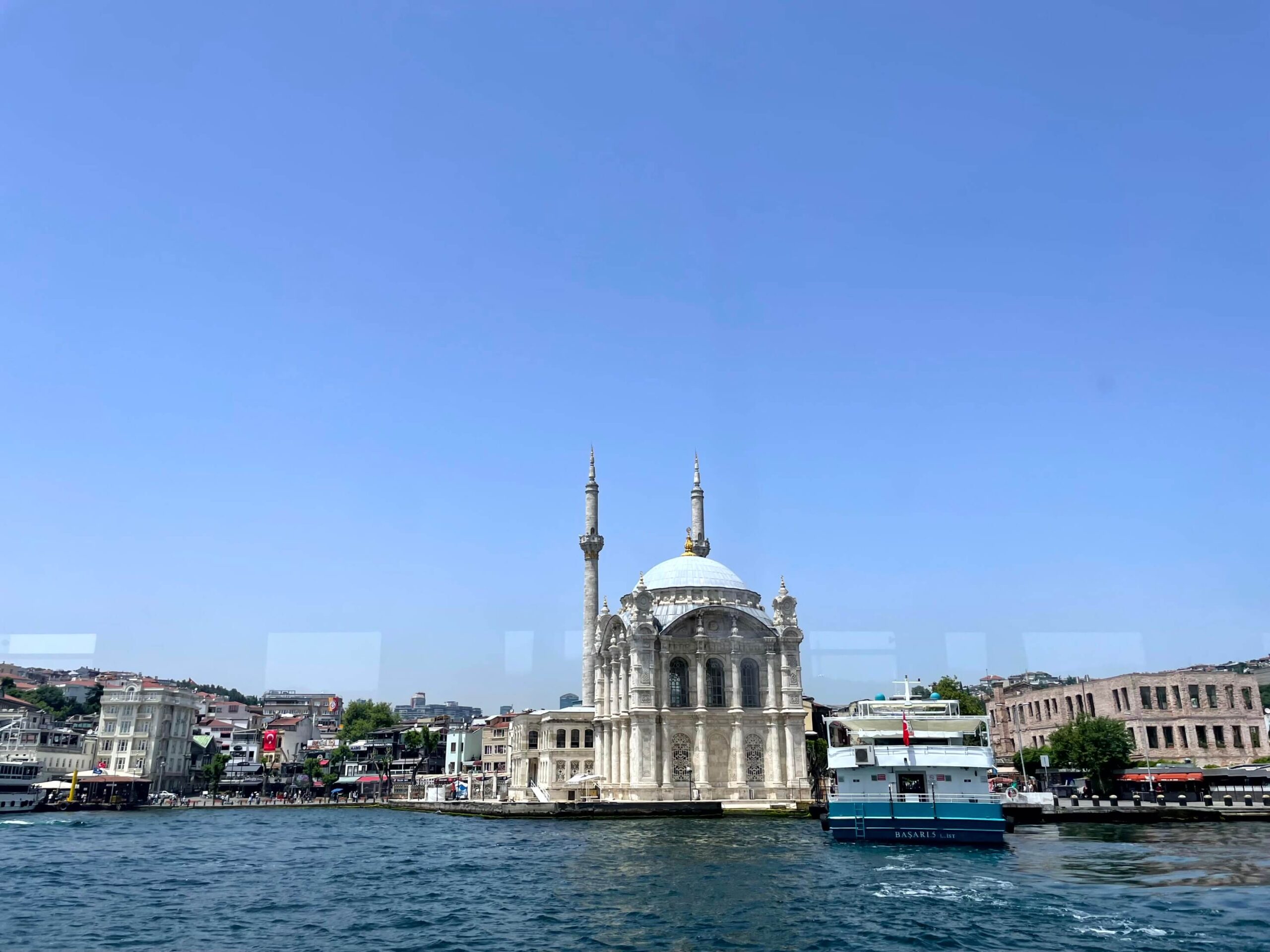 Ortaköy Mosque sits beside the Bosphorus with its domed roof and two minarets rising against a bright blue sky. Boats float near the shore as the mosque is seen from the water.