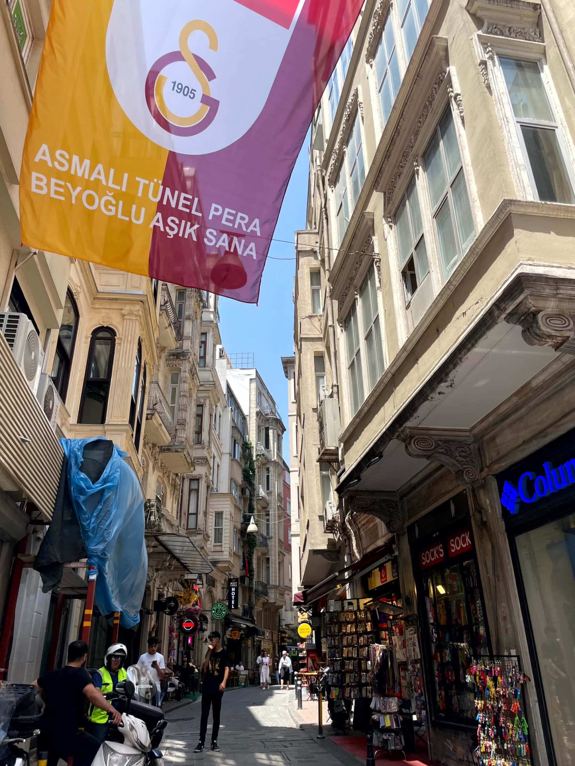 A narrow side street off İstiklal Street between tall historic buildings with shops selling tourist souvenirs on the bottom level. A large Galatasaray banner overhead reads "Asmalı Tünel Pera Beyoğlu aşık sana", a Turkish football chant.