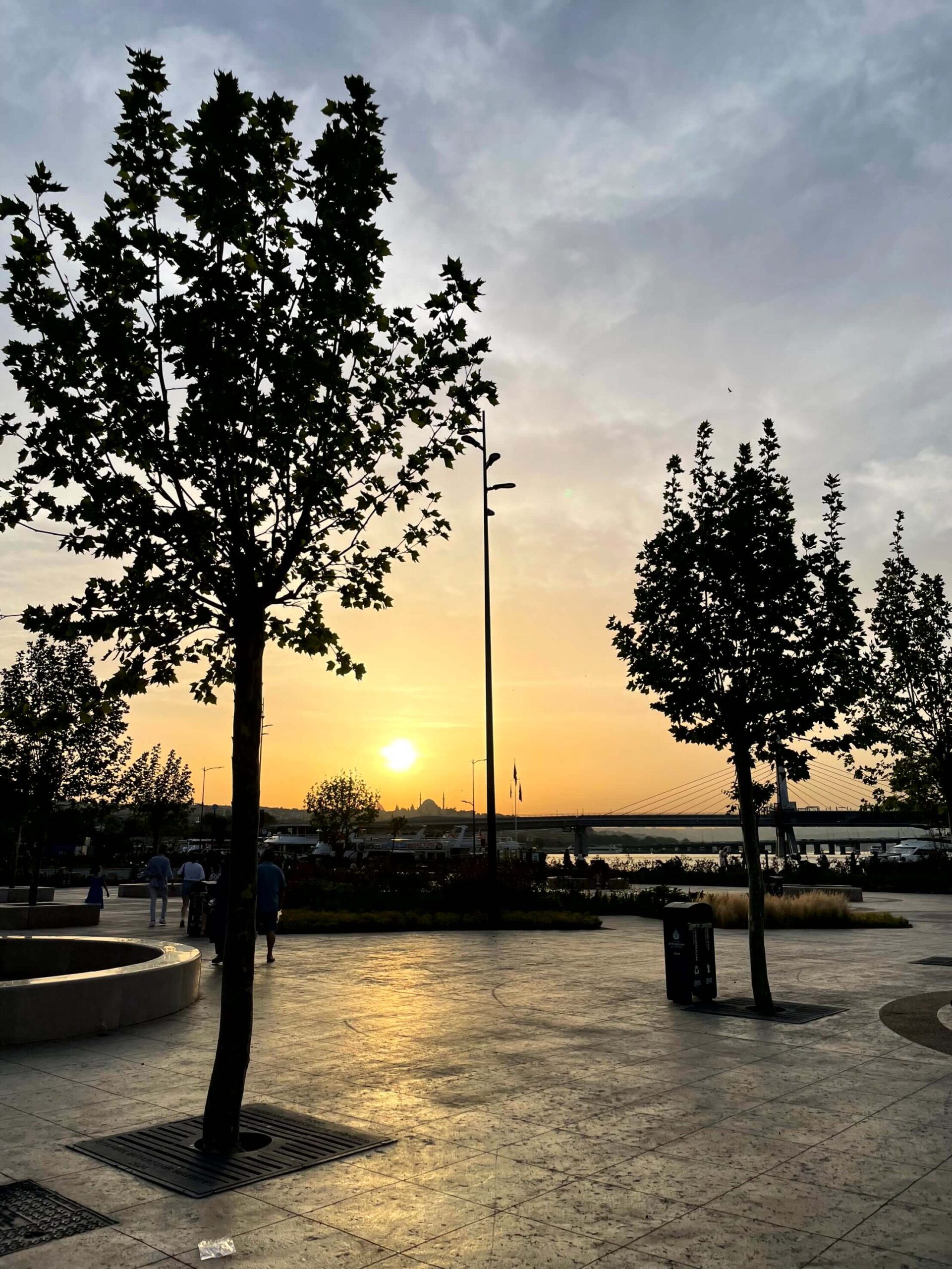Sunset over the Eminönü waterfront with trees silhouetted against a golden sky. The paved walking area reflects the evening light with the water and bridge in the background. A mosque is visible in the distance.
