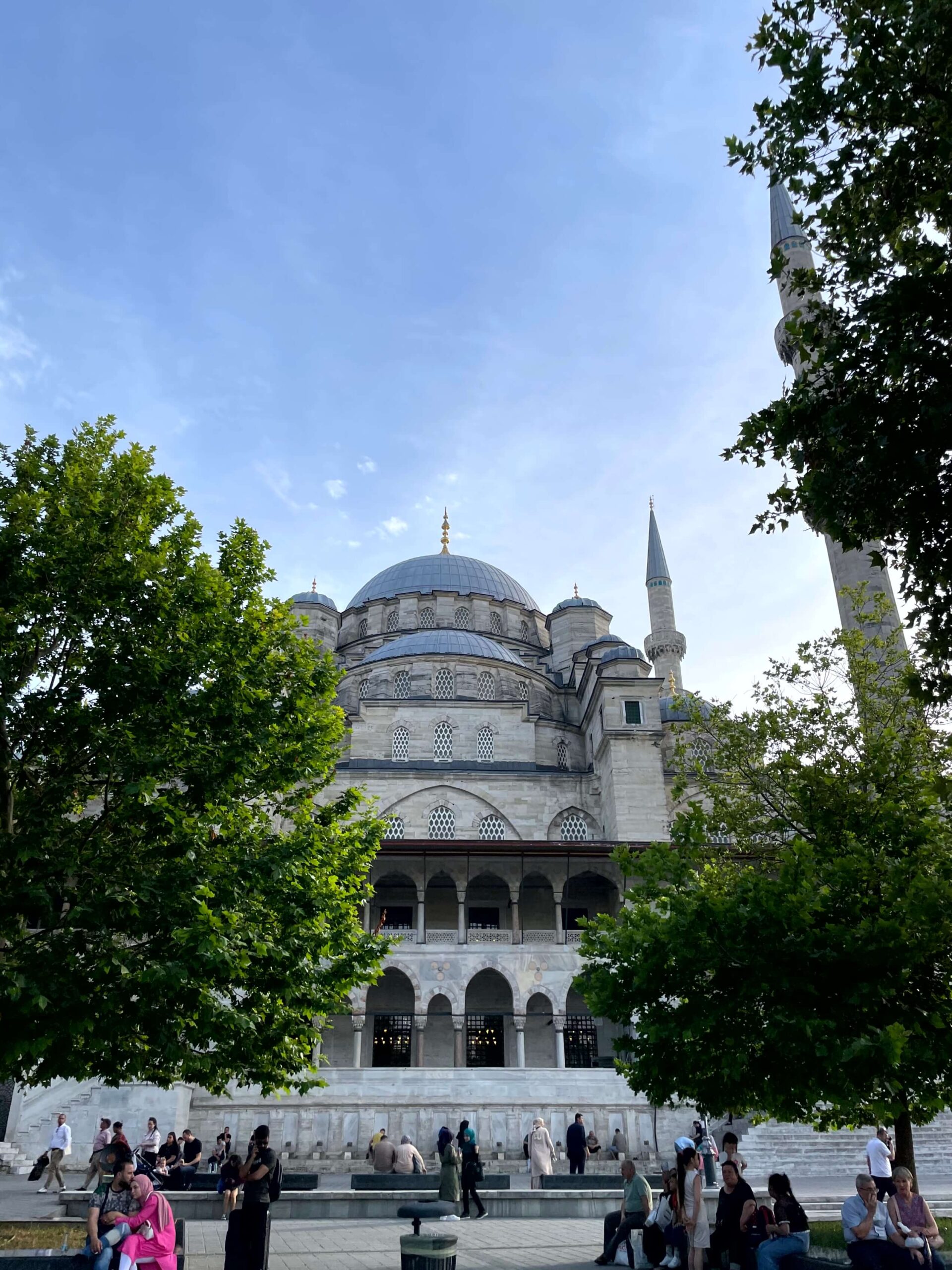 Yeni Camii (New Mosque) in Sirkeci framed by leafy green trees with its domes, arched windows, and minarets rising against a pale blue sky. People sit and walk through the open courtyard in front of the mosque.
