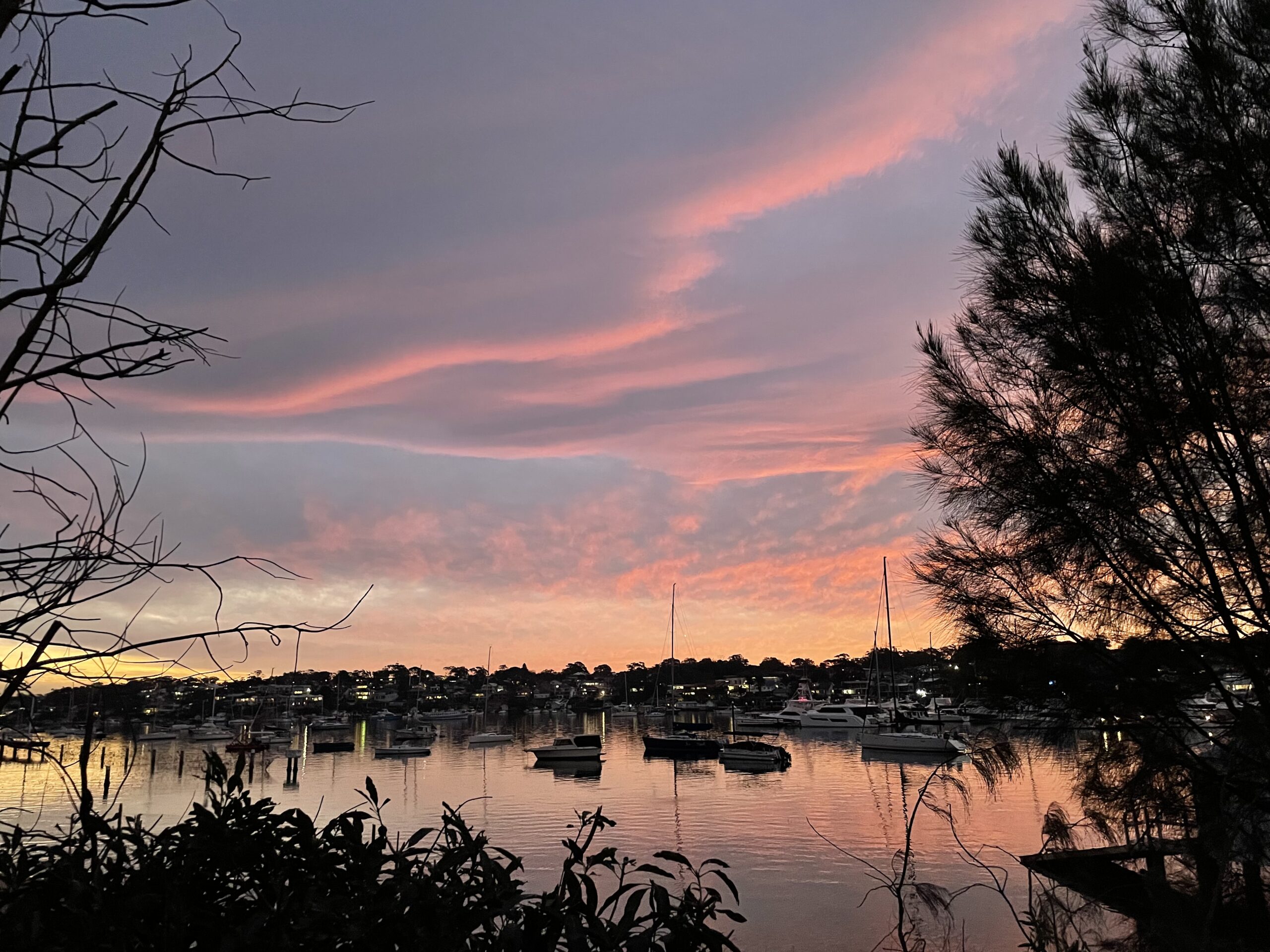 A vivid pink and orange sunset reflecting on the calm water of Gunnamatta Bay with small sail boats in the water.